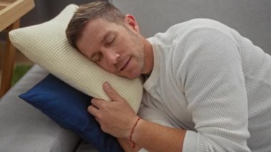 Rahat oturma odasındaki kanepede rahat bir şekilde uyuyan beyaz süveter giyen mavi yastıkla yumuşak yastığa sarılan bir adam..