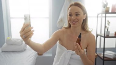 Görüntü sırasında elinde yağ şişesiyle gülümseyen bir kadın havluları ve rafları olan parlak bir kaplıca odasında arıyor..