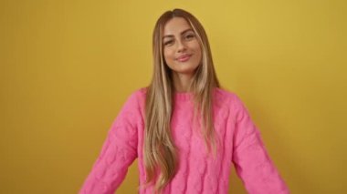 Sarışın bir kadının elleri tarafından yapılan kalp sembolü, izole edilmiş sarı arka planda romantik bir konsepte gülümsüyor, süveter giyiyor ve güzel bir şekilde duruyor.