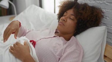 Sıcak bir yatak odasında kıvırcık saçlarıyla yatakta dinlenen yumuşak beyaz yatak ve doğal ışıklandırmayla ev ortamında huzurlu bir dinlenme ve rahatlık sergileyen bir kadın..