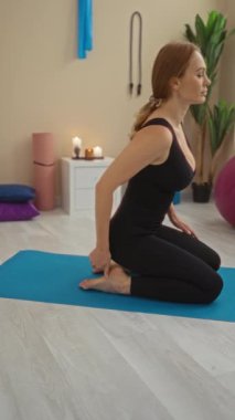 Evde yoga yapan bir kadın, sakin bir spor salonunda mavi bir paspasın üzerinde, bitkiler ve mumlarla huzurlu bir ortam yaratıyor..
