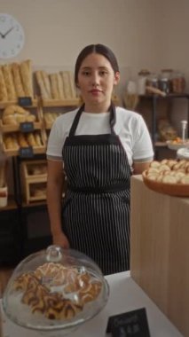 Çizgili önlüklü İspanyol kadın fırın işçisi arka planda ekmek ve hamur işleri sergilenen kapalı bir dükkanda kolları çapraz duruyor..