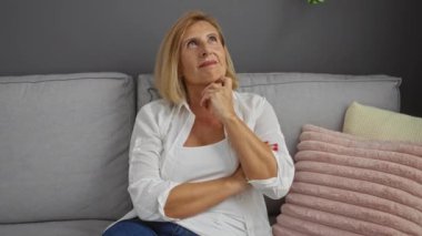 Rahat bir oturma odasında, kanepede oturmuş, beyaz bluz ve mavi kot pantolon, sarı saç, rahat iç mekan, olgun bir güzellik sergileyen bir kadın.