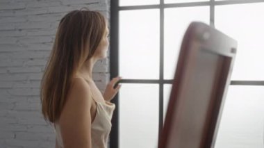 Yatak odasında durmuş, pencereden dışarı derin derin bakan, ışık saçan, sakin iç mekan ve sıcak ortam gösteren bir kadın.