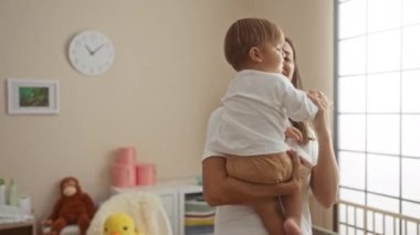 Kadın oğluyla neşeyle dans ediyor, parlak bir yatak odasında, yumuşak oyuncaklar ve mobilyalarla çevrili, sıcak ve sevgi dolu bir atmosfer yaratıyor..