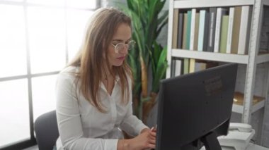 Modern bir ofiste bilgisayar ve kitaplıkla çalışan bir kadın, doğal ışık ve yeşillikle profesyonel içeriyi gösteren, genç ve çekici kadın profesyonel.