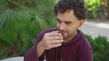 Genç adam şehir parkında stresli görünüyor. Etrafı yeşillikle çevrili, bordo kapüşonlu bir kapüşonlu giyiyor. Açık havada bir yorgunluk anı yaşıyor..