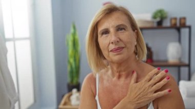 Kısa sarı saçlarıyla gülümseyen olgun bir kadın. Sağlık spa odasında, iç dekorasyonu ve parlak ışıklarıyla.