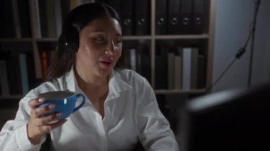 Ofiste elleri boş konuşan bir kadın elinde mavi bir kupayla arka planda kitaplıklar olan profesyonel bir iç mekanda nişanını sergiliyor.