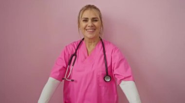 Steteskopla gülümseyen pembe önlüklü bir kadın izole edilmiş pembe arka planda kalbi oluşturur.