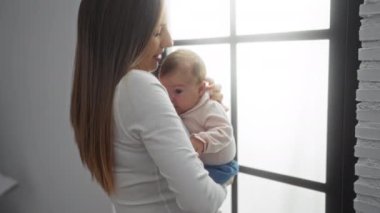 Kadın bebeği rahat bir oturma odasında kucağında tutuyor. Evin iç tarafındaki pencerenin önünde huzurlu anne-çocuk bağı sergiliyor..