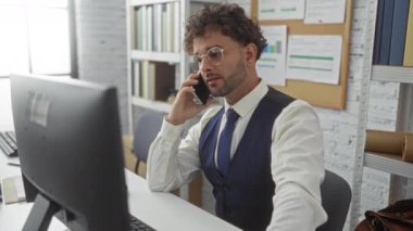 Ofisteki genç adam, profesyonel iç mekanlarda bilgisayar başında çalışırken telefonla konuşuyor..