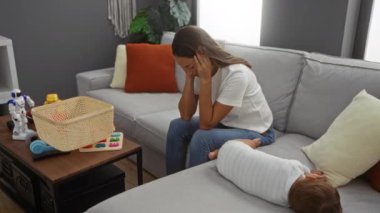 Bebek oyuncaklarla ve dekorlarla dolu modern bir oturma odasında uyurken ve aile dinamiklerini ve günlük yaşantısını resmederken bir kadın hayal kırıklığına uğramış bir ifadeyle koltukta oturuyor..