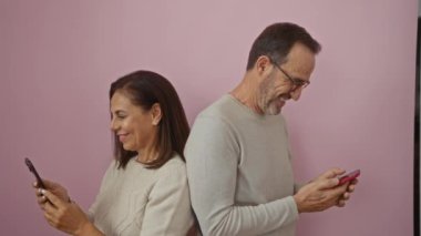 Aşkı ve iletişimi tasvir eden izole edilmiş pembe arka plana karşı telefonlarıyla gülümseyen orta yaşlı İspanyol çift.