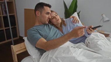 Yatak odasında rahat bir şekilde televizyon seyreden çift, yatakta sarılırken gülümseyen erkek ve kadın, evde sıcak ve samimi bir atmosfer yaratıyor..