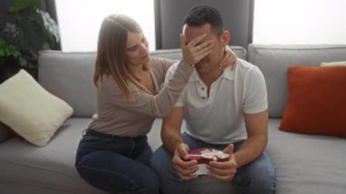 Oturma odasında oturan bir çift, hediyeli sürpriz bir kadın, ikisi de gülümsüyor, evde hassas bir anın tadını çıkarıyor..