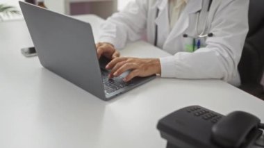 Klinikteki genç erkek doktor, boynunda steteskopla dizüstü bilgisayar kullanıyor. Parlak bir muayenehanede çalışırken gülümsüyor..