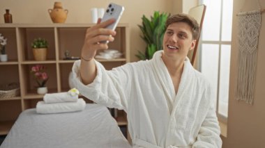 Beyaz bir bornoz giyerken, spa odasında selfie çeken ve mutlu bir şekilde gülümseyen genç bir adam. Etrafı zenginlik dekoru ve iç mekan bitkileriyle çevrili.