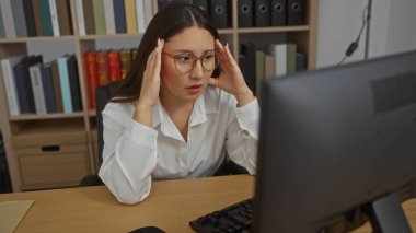 Ofis masasında çalışan, gözlüklü, başı kitaplıklarla çevrili stresli bir kadın.