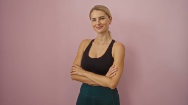 Pembe bir duvara karşı güvenle poz veren bir kadın, güzel ve formda bir gülümseme ve kollarını kavuşturmuş..