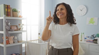 Yatak odasındaki İspanyol kadın, yumuşak oyuncaklar ve ev dekoruyla çevrili bir bebek beşiğinin yanında sıcak ve huzurlu bir atmosfer yaratarak bir fikir jesti yaparken gülümsüyor..