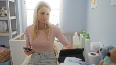 Konforlu yatak odasında telefon ve tablet kullanan bir kadın. Masada bebek eşyaları var. Modern evdeki çok görevli yaşam tarzını gösteriyor..