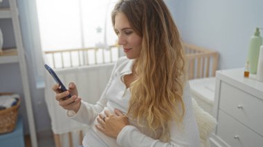 Yatak odasında rahat bir şekilde uzanan, hafif ışıklandırmalı akıllı bir telefon tutan bir kadın. Arka planda modern dekorasyon ve mobilyalar eşliğinde bir beşik görülüyor..