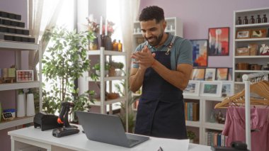 Delikanlı, elinde dizüstü bilgisayarla tezgahın arkasında dikilip el çırpıyor. Etrafı bitkilerle ve çeşitli dekorasyonlarla çevrili..