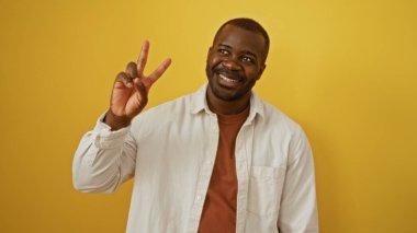 Genç, yakışıklı, siyah, izole edilmiş sarı bir arka planda gülümseyen, günlük, şık kıyafetler giyen, barış işareti yapan, pozitif enerji ve güven saçan bir adam..