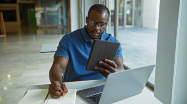 Genç, yakışıklı, Afrikalı, Amerikalı, tablet üzerinde çalışan ve bir ofis ortamında dizüstü bilgisayarla notlar yazan bir adam..