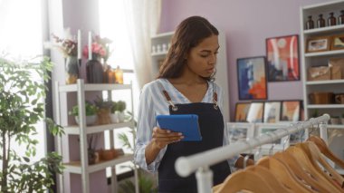 Önlüklü genç bir kadın, arka planda bitki ve sanat eserleri olan ev dekorasyon dükkanında tablet okuyor.