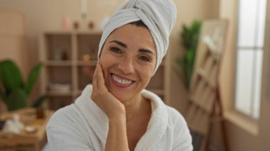 Kafasında bornoz ve havlu olan bir güzellik merkezine gülümseyen bir kadın, rahat bir sağlık merkezinde sergilenen orta yaşlı, dingin, İspanyol bir kadının dinlenme zamanının tadını çıkardığını gösteriyor..