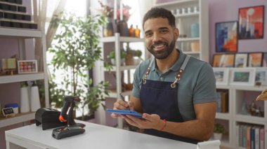 Kapalı bir dekor dükkanında çalışan genç bir adam elinde tabletle gülümsüyor, etrafı dekorasyon malzemeleri ve bitkilerle çevrili iyi aydınlatılmış bir dükkanın içinde.