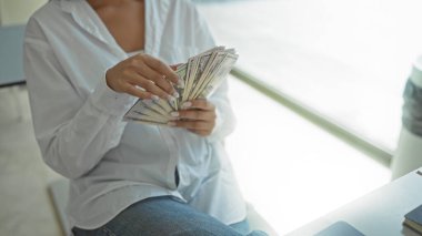 Günümüz ofis ortamında bize dolar sayan genç bir kadın. Büyük pencerelerden içeri güneş ışığı giriyor.