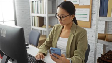Ofiste çalışan genç Çinli kadın, akıllı telefon ve bilgisayar kullanıyor, görevlere odaklanmış, modern kapalı iş yeri, profesyonel ve üretken çevre.