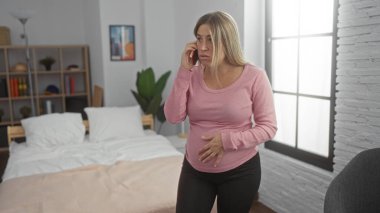 Hamile kadın rahat bir yatak odasında yatağında, elleri karnının üstünde, telefonla konuşuyor, pencereden yansıyan güneş ışığı huzurlu bir ev ortamında beklediği figürü vurguluyor..