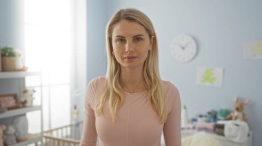 Yatak odasında durmakta olan, yumuşak ışıklandırmalı, sarı saçlı, bebek beşiği ve kişisel dekorları olan, sakin ve besleyici bir atmosferi olan bir kadın..