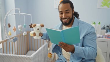 Bebek odasında kitap okurken gülümseyen bir adam elinde beşikteki oyuncakla pencereden içeri doğru hafif bir ışık süzülüyor.