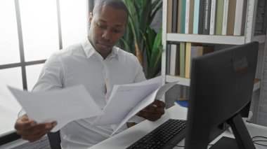 Evraklar ve bilgisayarla dolu bir ofiste çalışan, işine odaklanmış, resmi tişört giyen, modern iş yerinde profesyonel ve çekici tavırlar sergileyen genç bir adam..