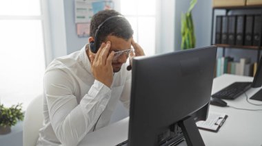 Kulaklık takan ve gözlük takan İspanyol genç adam stresli görünüyor. Modern ofis ortamında bilgisayar monitörü ve ofis malzemeleriyle dolu bir masada oturuyor..