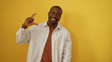 Beyaz gömlekli genç adam parmaklarıyla büyük jest yapıyor. Parlak sarı duvara yaslanıyor. Pozitif ve kendine güvenini gösteriyor..