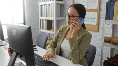 Genç Asyalı kadın telefonda konuşuyor ve ofis ortamında bilgisayar kullanıyor. Profesyonel bir çevreyi yansıtıyor..