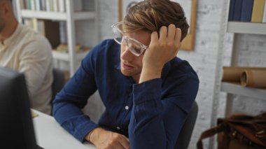 Ofiste çalışan ve masasında yorgun ve stresli görünen erkekler kapalı alanda yoğun bir iş ortamını ön plana çıkarıyorlar..