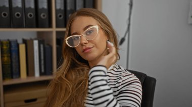 Ofiste güven içinde poz veren, gözlüklü, çizgili tişörtlü, kitaplık ve klasörlerle çevrili bir kadın..