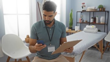 Klinik odasında elinde bir pano olan genç bir adam telefonuna bakıyor, muhtemelen hasta bilgilerini belgeliyor ya da gözden geçiriyor, iyi aydınlatılmış bir ortamda masaj masasının yanında oturuyor..