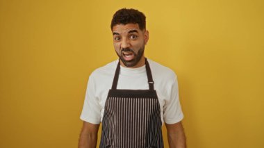 Beyaz tişörtlü ve çizgili önlüklü İspanyol genç adam izole edilmiş sarı arka plan üzerinde anlamlı bir yüz yapıyor.