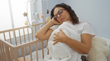 Kadın, beşiğin yanındaki rahat yatak odasında huzur içinde dinleniyor. Yumuşak bir iç mekânda sıcaklığını, huzurunu ve annelik bağını ifade ediyor..