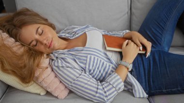 Kanepede uyuyan genç bir kadın rahat oturma odasında kitap tutuyor.