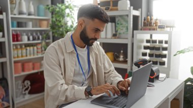 Genç, İspanyol, sakallı bir adam ev dekorasyonu mağazasında dizüstü bilgisayar üzerinde çalışıyor, çeşitli dekoratif eşyalar, raflar ve kapalı alandaki bitkilerle çevrili..