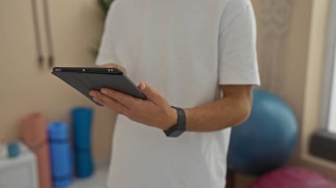 Spor salonundaki adam spor ortamındaki teknolojik etkileşimi gösteren egzersiz ekipmanlarıyla tablet tutuyor, günlük beyaz gömlek giyiyor..